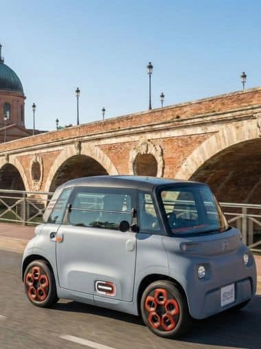 Une voiture électrique sans permis Citroën Ami grise avec des enjoliveurs orange, circulant à Toulouse devant le Pont-Neuf en briques roses.