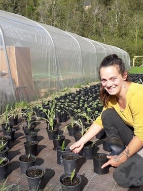 L'agricultrice Célia Muñoz, fondatrice de l’association Belles à croquer s'occupe avec le sourire de plants dans une pépinière. Elle est agenouillée sur le sol, entourée de rangées de jeunes plants dans des pots. À sa gauche, une serre en tunnel est visible.